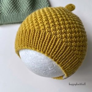 May include: A mustard yellow knitted beanie with a textured stitch pattern and a ribbed brim, featuring a small top knot and a tag with the text happyknitball. A sage green knitted beanie is partially visible in the upper left corner.