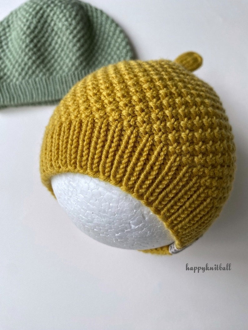 May include: A mustard yellow knitted beanie with a textured stitch pattern and a ribbed brim, featuring a small top knot and a tag with the text happyknitball. A sage green knitted beanie is partially visible in the upper left corner.