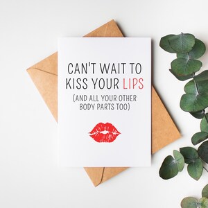 May include: A white greeting card with the text "CAN'T WAIT TO KISS YOUR LIPS (AND ALL YOUR OTHER BODY PARTS TOO)" and a red lip print. The card is on a brown envelope, with green leaves in the background.