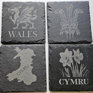May include: A set of four square slate coasters, each with a different engraved design. Designs include a Welsh dragon, a crown with feathers, a map of Wales, and daffodils. The words "WALES" and "CYMRU" are also engraved.
