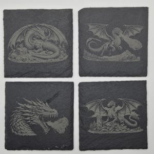 May include: A set of four square slate coasters, each featuring a different dragon design. The dragons are etched onto the dark grey slate and shown in various poses, including a dragon resting, flying, breathing fire, and near a castle.