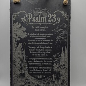 May include: A rectangular black slate plaque featuring an engraved illustration of a shepherd with sheep and the full text of Psalm 23. The plaque has a rough, natural edge and includes a thick, tan-coloured jute rope attached for hanging.