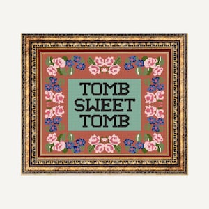 Op de afbeelding: Een ingelijst kruissteek met een bloemige rand en de tekst "TOMB SWEET TOMB" in zwarte letters op een teal achtergrond.