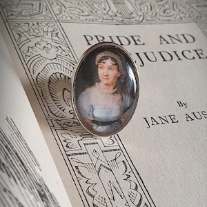 Könnte beinhalten: Ein silberner Ring mit einem Glas-Cameo, das ein Porträt von Jane Austen zeigt, der Autorin von "Pride and Prejudice". Der Ring liegt auf einem Buch mit dem Titel "Pride and Prejudice" und dem Namen der Autorin, "Jane Austen".