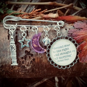 Peut inclure: Épingle de sûreté argentée avec des breloques pendantes. Les breloques comprennent une réplique de Big Ben, une étoile, une lune violette et un pendentif circulaire avec la citation "Second star to the right and straight on 'til morning" de Peter Pan.