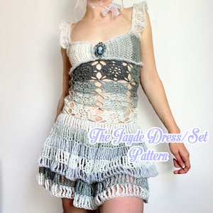 May include: A crocheted dress and matching top set in white and gray with a ruffled skirt. The dress has a cameo brooch and the text "The Jayde Dress/Set Pattern" is printed on the skirt.