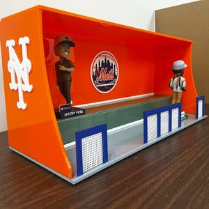 Pode incluir: Uma vitrine laranja e branca com duas figuras de baseball. A vitrine tem um logotipo dos Mets e o nome "Jeremy Peña" em um pequeno sinal. A vitrine tem uma base cinza e cercas azuis e brancas.