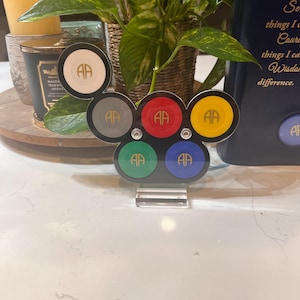 May include: A set of six round, colorful, decorative coasters with the letters "AA" in gold lettering on each coaster. The coasters are arranged in a circle on a black stand with a clear acrylic base.