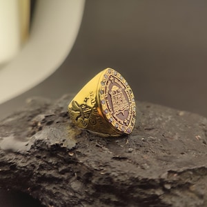 May include: A gold-colored ring with a shield-shaped face. The ring features a detailed crest with text and a border of small, dark stones. The band of the ring has additional engraved details. The ring is displayed on a dark, textured surface.