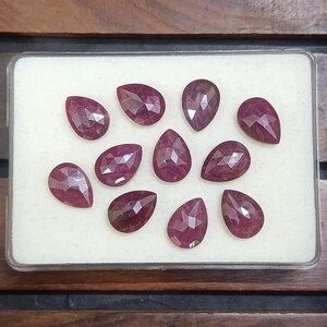 Puede incluir: Colección de diez rubíes facetados en forma de pera. Las piedras preciosas son de un color rojo intenso y rico y se exhiben en una caja de plástico transparente con un interior blanco. Los rubíes están dispuestos en un patrón ordenado.