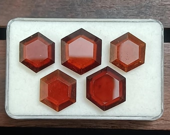 May include: Five faceted, hexagonal gemstones in a clear plastic case. The gems are a deep reddish-brown color, with a polished, reflective surface. The case sits on a wooden surface.