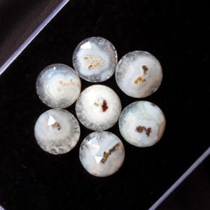 Può includere: Sette gemme rotonde e lucide, con una combinazione di colori bianco e grigio, disposte a cerchio. Ogni pietra presenta motivi interni marroni e bianchi unici e una superficie sfaccettata. Le gemme sono esposte su una superficie nera.