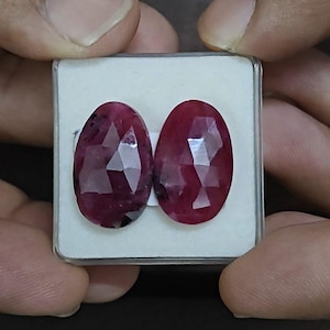 May include: Two oval-shaped, faceted red gemstones with a deep crimson hue and reflective surfaces. The stones are set side-by-side on a white foam insert within a clear, square plastic display case, showcasing their polished, multi-faceted cut.