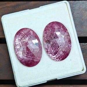 Puede incluir: Dos piedras preciosas ovaladas y facetadas de color rojo intenso. Las gemas se exhiben sobre una superficie blanca, mostrando sus intrincados patrones internos y superficies reflectantes. Las piedras preciosas son probablemente para la fabricación de joyas.