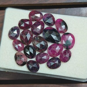 May include: A collection of fifteen oval-shaped, faceted gemstones in shades of deep magenta, dark pink, and black. Each stone features a rose-cut surface with geometric facets that reflect light, arranged in a cluster on a white foam display pad.