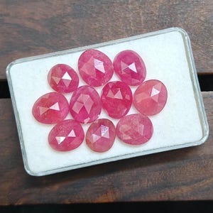 May include: A collection of nine oval-shaped, faceted pink rubies arranged on a white foam insert within a rectangular display case. Each gemstone features a polished surface with geometric facets that reflect light, showcasing a vibrant pink hue.