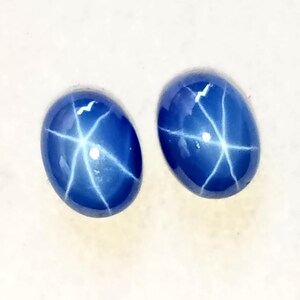 Peut inclure: Deux pierres précieuses ovales bleu foncé avec un motif en forme d'étoile. L'effet d'étoile est créé par des lignes blanches rayonnant du centre de chaque pierre. Les gemmes ont une surface polie et brillante.