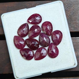 May include: Ten faceted, pear-shaped rubies in a clear plastic container. The gemstones are a deep red colour. The cut of the rubies is designed to maximise light reflection. The rubies are displayed on a white surface.