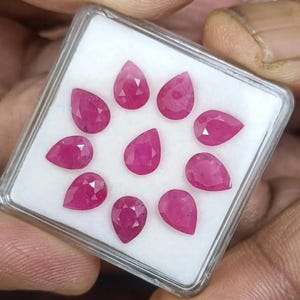 Op de afbeelding: Een verzameling van tien peervormige, gefacetteerde robijnen, gerangschikt in een transparante plastic doos. De edelstenen hebben een levendige, dieprode kleur en worden gepresenteerd tegen een witte achtergrond. De doos wordt in een hand gehouden.