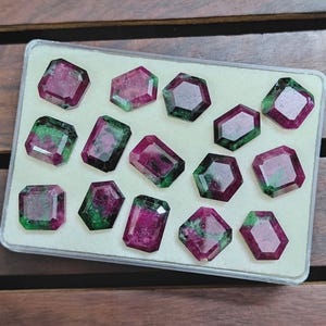 May include: A collection of faceted gemstones in a clear plastic case. The gems feature a mix of deep red and green hues, with various geometric shapes including rectangles and octagons. The gems are displayed on a white surface.