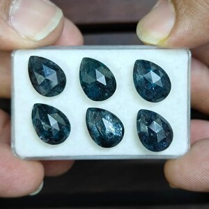 Op de afbeelding: Zes peervormige, gefacetteerde edelstenen in een witte doos. De diepblauwe tot teal-kleurige stenen hebben facetten die het licht vangen. De doos wordt door handen vastgehouden, waardoor de vorm en kleur van de stenen worden benadrukt.