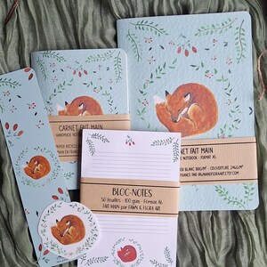 Peut inclure: Ensemble de papeterie fait main avec un motif de renard endormi. L'ensemble comprend des carnets, un marque-page et un bloc-notes, tous sur fond bleu clair avec des éléments botaniques. On peut lire "CARNET FAIT MAIN" et "BLOC-NOTES".