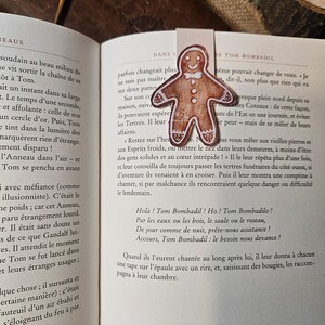 Op de afbeelding: Een bladwijzer in de vorm van een peperkoekmannetje met bruin glazuur, witte details en een strikje, geplaatst tussen de pagina's van een open boek met Franse tekst. De bladwijzer rust tegen het bedrukte papier van het boek aan.