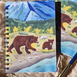 Puede incluir: Un cuaderno de espiral con una ilustración pintada de un oso pardo y su cría caminando por la orilla de un río. El fondo muestra un bosque verde, árboles amarillos y un gran pico de montaña azul bajo un cielo despejado, con colores vivos.