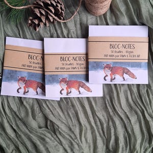 May include: Three "Bloc-Notes" notepads with a watercolor illustration of a fox in a snowy landscape. Each notepad has a brown paper band with text in French. The notepads are on a green fabric surface, with a pinecone and twine in the background.