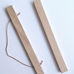 Pode incluir: Dois cabides de cartaz de madeira de cor clara com um acabamento natural. Cada cabide é um retângulo longo e fino com um pequeno orifício em cada extremidade. Uma corda castanha está presa a um dos cabides. Os cabides estão sobre uma superfície branca.