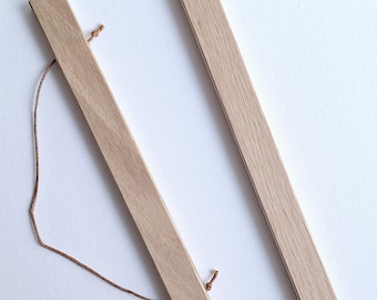 May include: Two light-colored wooden poster hangers with a natural finish. Each hanger is a long, thin rectangle with a small hole at each end. A brown string is attached to one of the hangers. The hangers are lying on a white surface.