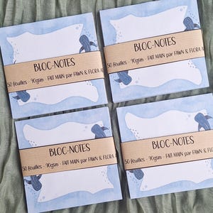 Peut inclure: Quatre blocs-notes carrés avec une bordure bleu clair et une illustration de baleine. Chaque bloc-notes porte une bande marron avec le texte "BLOC-NOTES" et "50 feuilles - 90gsm - FAIT MAIN par FAWN & FLORA A".