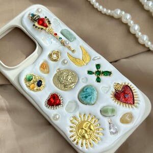 May include: A white phone case adorned with various decorative elements. It features a jeweled wand, gold wings, a sun, a coin, and a cross. The case is embellished with red, green, and blue gemstones, pearls, and other decorative accents.