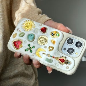 Può includere: Custodia per telefono bianca ornata con ciondoli dorati a forma di sole, moneta e ala, insieme a pietre preziose rosse, verdi e blu. La custodia presenta anche una bacchetta con punta di gioiello e un gruppo di perle.