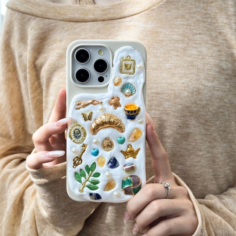 Op de afbeelding: Witte iPhone-hoes versierd met gouden, blauwe en groene versieringen. De hoes heeft een verscheidenheid aan bedels, waaronder een halve maan, een sleutel, een vlinder en een klein kopje.