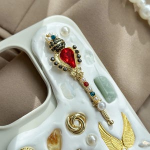 May include: A white phone case with a decorative design featuring a gold scepter with a red gem, pearls, and colorful jewels. Additional gold and pearl embellishments include wings and a spiral.