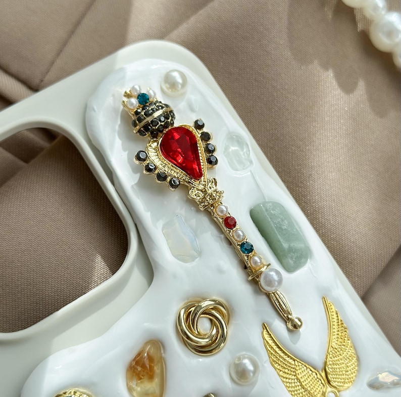 May include: A white phone case with a decorative design featuring a gold scepter with a red gem, pearls, and colorful jewels. Additional gold and pearl embellishments include wings and a spiral.