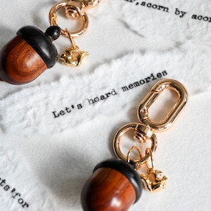 May include: Two acorn-shaped keychains with gold-colored hardware and a small squirrel charm. The acorns are brown with black caps. The text "Let's hoard memories" is printed on the white background.