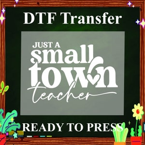 May include: A white iron-on transfer design on a black chalkboard background. The design says "Just a small town teacher" with a heart in the word "town". The text "DTF Transfer" is at the top of the chalkboard and "READY TO PRESS" is at the bottom.