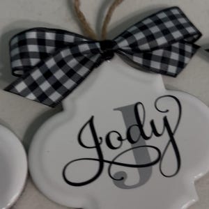 May include: White ceramic ornament with the name "Jody" in elegant script. The letter "J" is gray. A black and white checkered bow and a twine loop are attached for hanging. A decorative item.