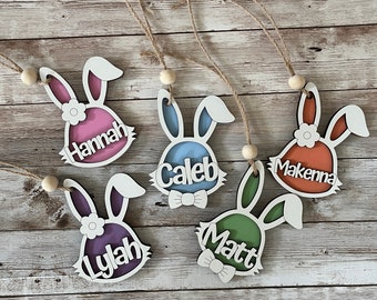 May include: Five wooden bunny-shaped ornaments with names written on them. The names are Hannah, Caleb, Makenna, Lylah, and Matt. The bunnies are painted in different colors, including pink, blue, orange, purple, and green.