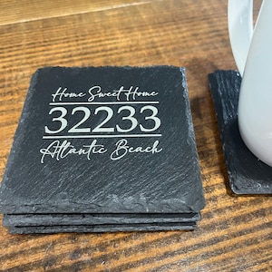 May include: A stack of black slate coasters with the text "Home Sweet Home 32233 Atlantic Beach" in white script. The coasters are rough-edged and sit on a wooden surface. A white ceramic pitcher is partially visible.