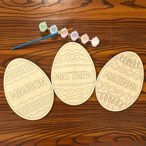 May include: Three wooden Easter eggs with painted designs and names "Mayson", "Matthew", and "Makenna". The eggs are on a wooden surface with paint pots and a paintbrush.