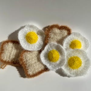 May include: A set of crocheted kitchen scrubbers shaped like toast and fried eggs. The scrubbers are made with white and brown yarn and have a yellow yarn centre.