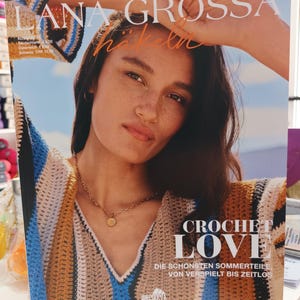 May include: A magazine cover featuring a woman wearing a crocheted top with a striped pattern in shades of brown, blue, and white. The magazine is titled "Lana Grossa Häkeln Crochet Love" and the text "Die schönsten Sommerteile von verspielt bis zeitlos" is printed below the title.