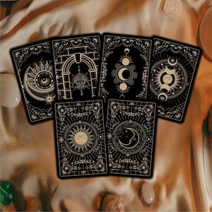 May include: A set of tarot cards with black backgrounds and gold celestial designs. Each card features unique illustrations of moons, suns, and stars, framed by ornate borders. The cards are arranged on a cream-coloured fabric.