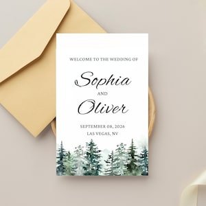May include: A wedding sign with the names Sophia and Oliver, the date September 08, 2026, and location Las Vegas, NV. The sign has a watercolor forest scene at the bottom and is accompanied by a tan envelope.
