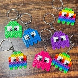 May include: Keychains featuring pixelated ghost-like figures in various colors, including green, blue, purple, pink, and rainbow. Each keychain has a silver ring and chain. The figures have black and white details, resembling classic video game characters.