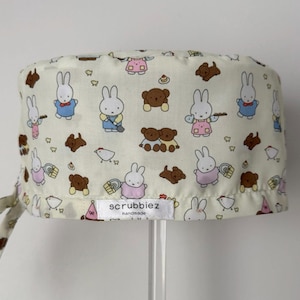 May include: A cream-coloured scrub cap with a repeating pattern of cartoon bunnies, teddy bears, and chickens. The cap has a tie closure and a label that reads "scrubbiez handmade."