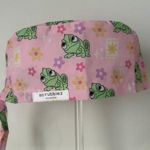 May include: Pink scrub cap patterned with green chameleons, yellow and purple flowers, and golden lanterns. The cap has a tie closure and a small white tag that reads "scrubbiez handmade".